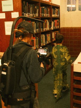 A Steadicam operator following a cast member in a dinosaur costume through a library