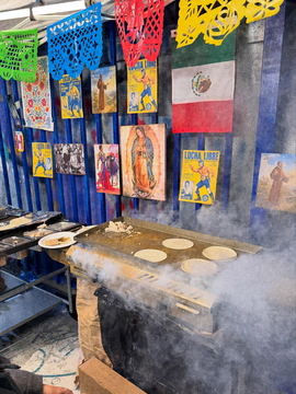 A market stall set hung with papel picado, a Mexican flag and devotional prints, smoke rising from the griddle